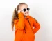 Dievčatko v oranžovej mikine so slnečnými okuliarmi na bielom pozadí.